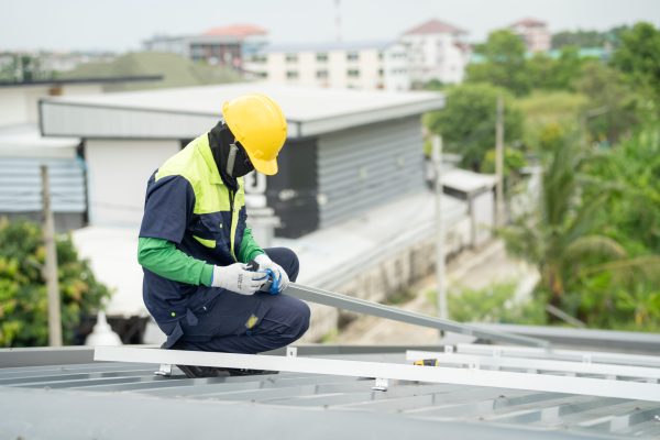 Builder mounting photovoltaic solar modules on roof of house. Back view of man engineer in helmet installing solar panel system outdoors. Concept of alternative and renewable energy.